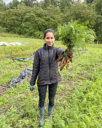 A student is standing in a field holding a bunch of carrots up to the camera. She is wearing a bl...